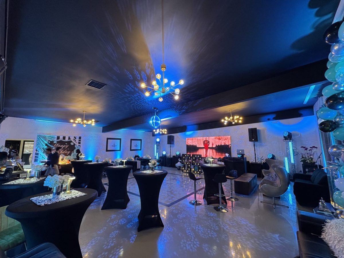 Main room set for a cocktail party with black high-top tables, gold chandeliers and blue uplighting