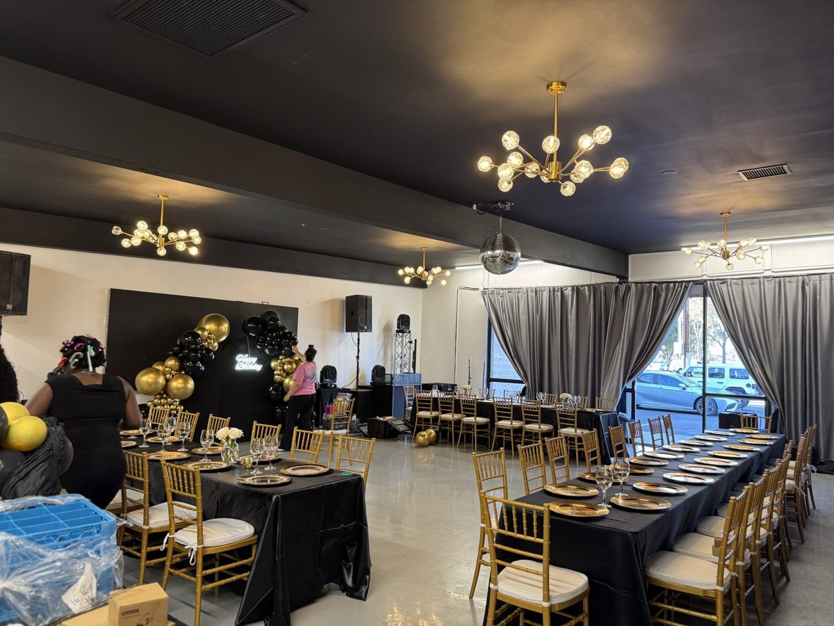 Banquet layout with long black-linen tables, gold charger plates and gold chiavari chairs in front of a black and gold balloon backdrop
