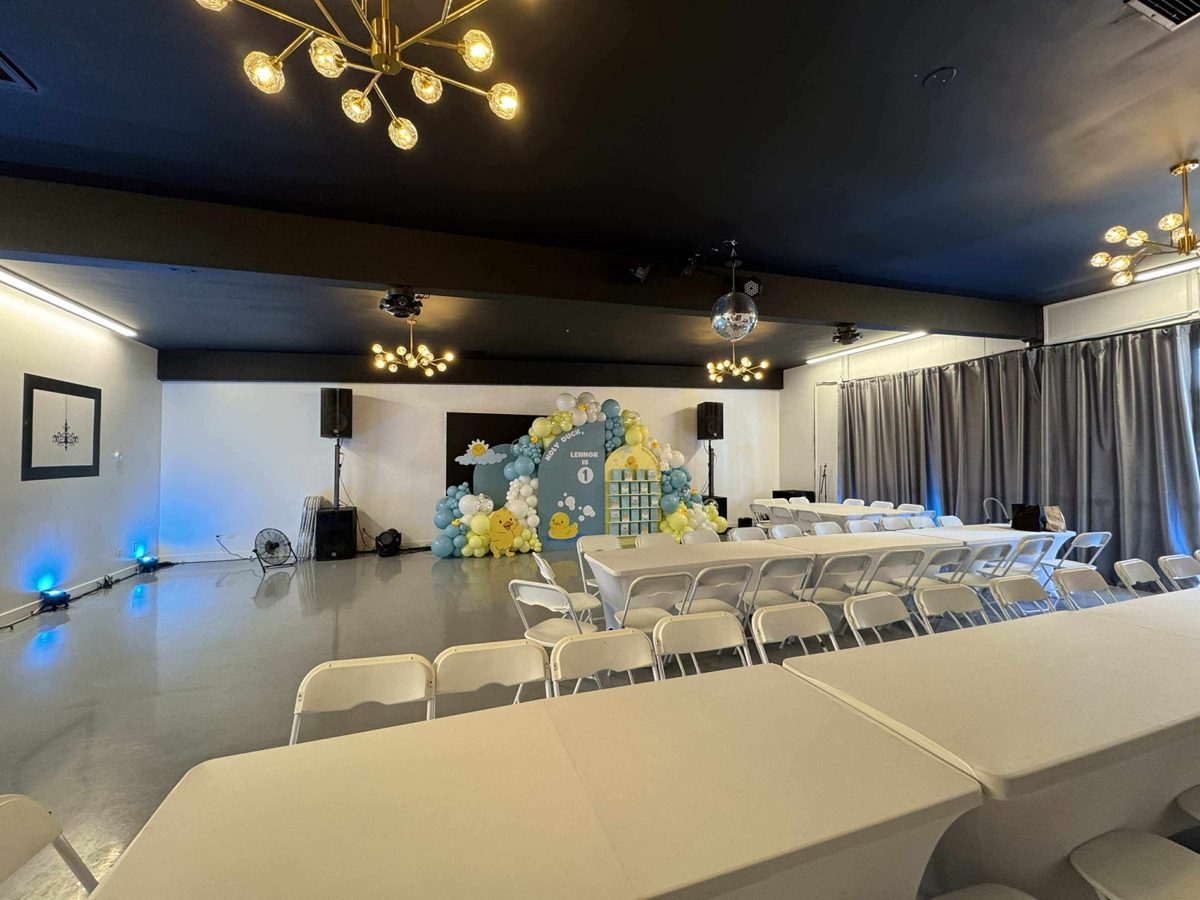 Main room with long white tables and white chairs facing a balloon backdrop, under gold chandeliers